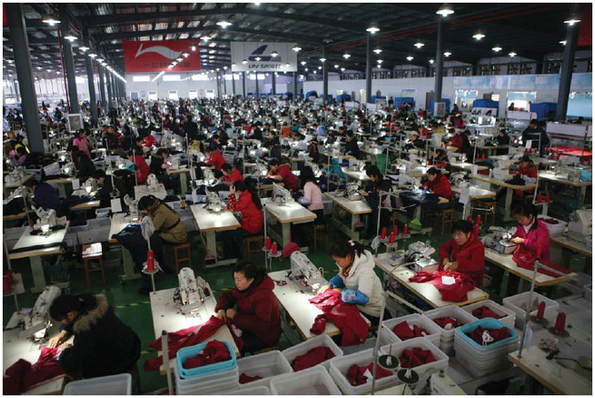 a photo of many people working in a sports clothing factory in China. there are many rows of sewing machines with a worker manning each one. The room is large, but still cramped with the amount of people working.