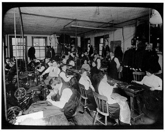 a black and white photo of many women working in the triangle shirtwaist factory. there are many rows of sewing machines with fabric strewn about the cramped room